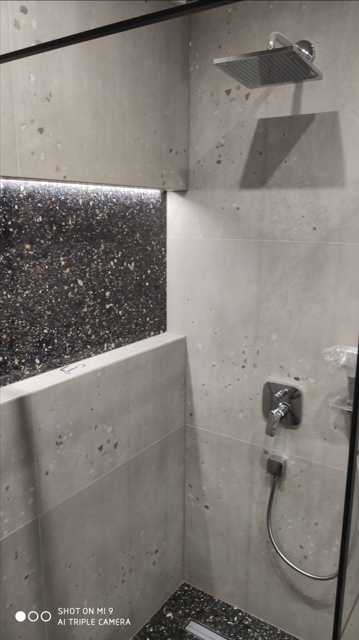 Nowoczesna kabina prysznicowa z szarymi płytkami imitującymi lastryko, czarną wnęką z oświetleniem LED i chromowaną armaturą.