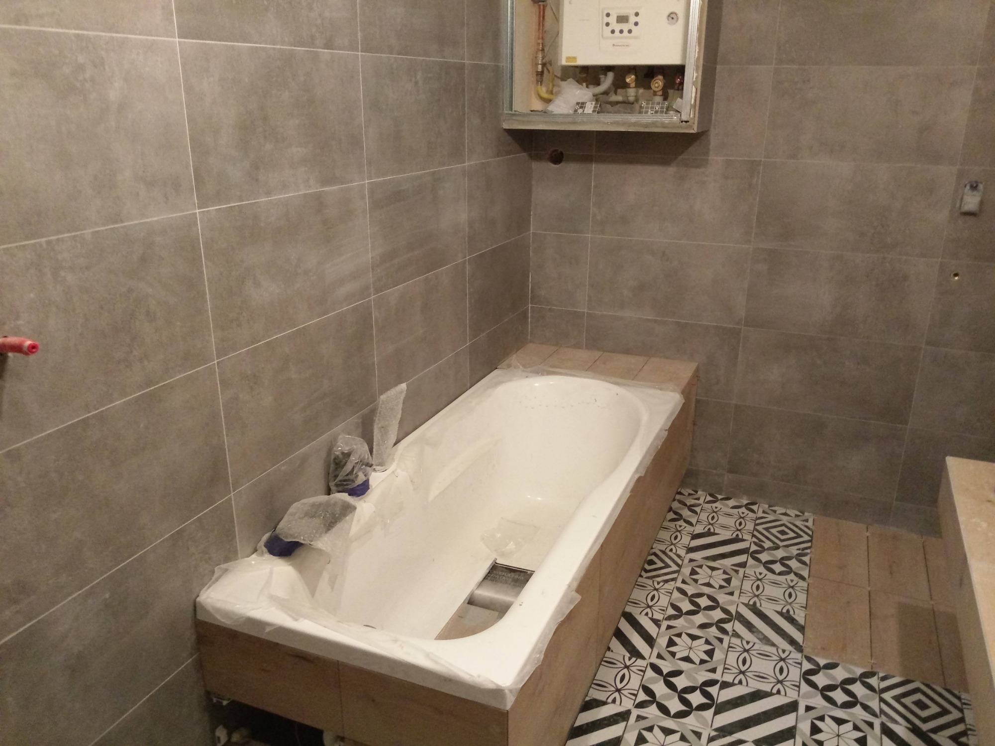 Wanna obudowana płytami, zabezpieczona folią, na tle szarych płytek ściennych i podłogi z wzorzystych płytek, widoczny fragment instalacji nad wanną.