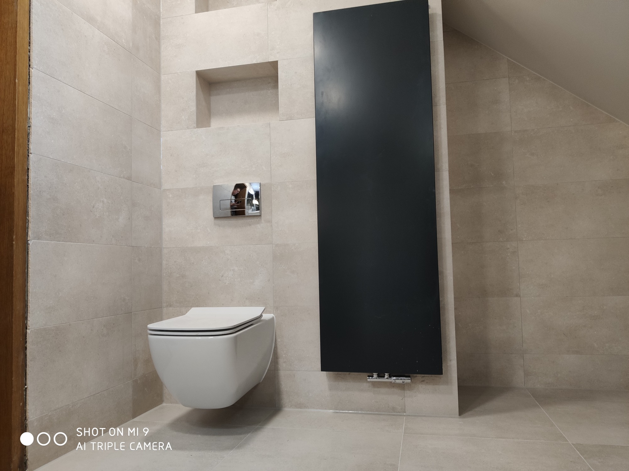 Nowoczesna łazienka na poddaszu z beżowymi płytkami, wiszącą toaletą, czarnym grzejnikiem panelowym i wnęką w ścianie.