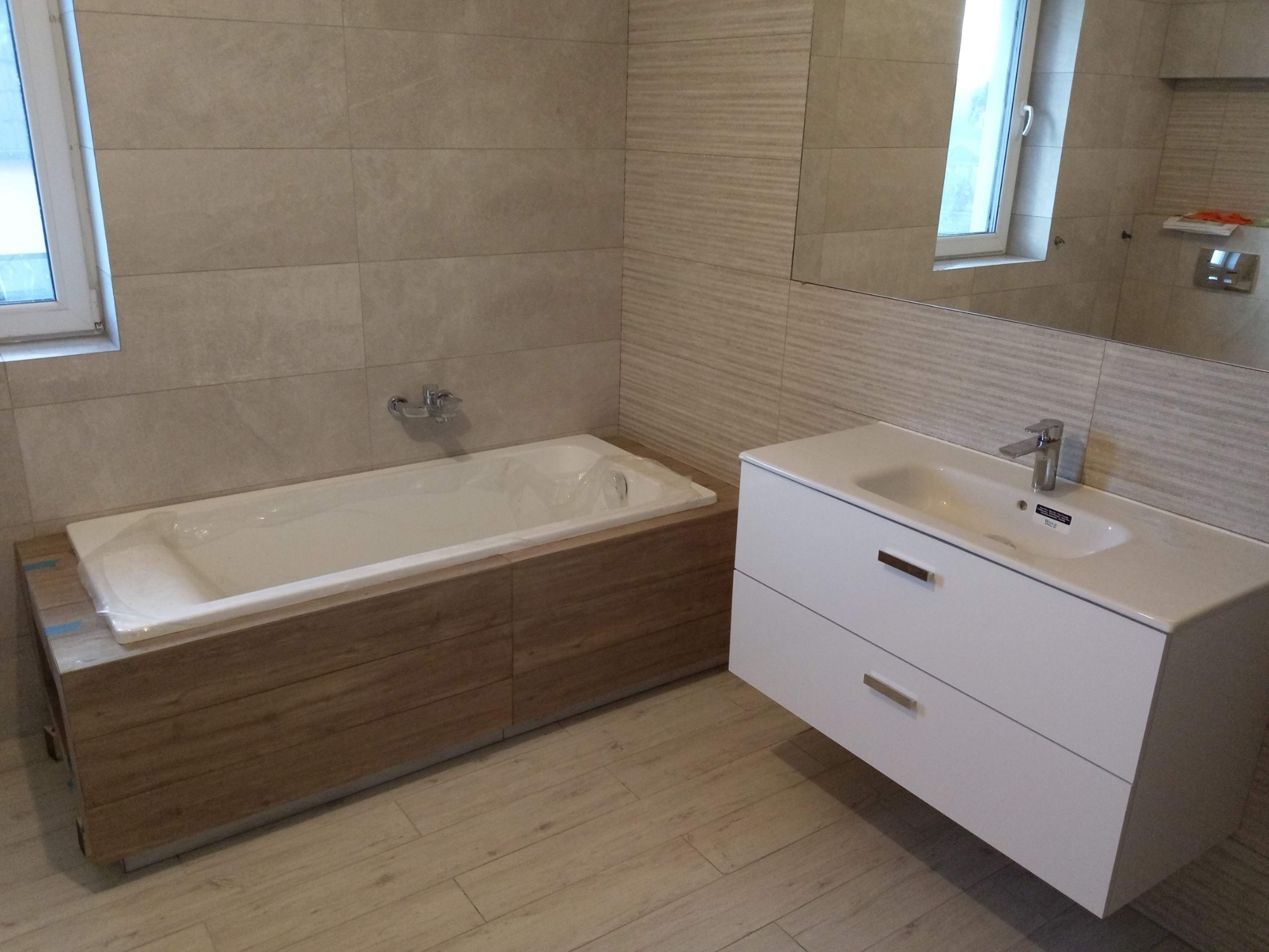 Nowoczesna łazienka z beżowymi płytkami, białą umywalką z szafką podwieszaną i obudowaną wanną, zabezpieczoną folią ochronną.