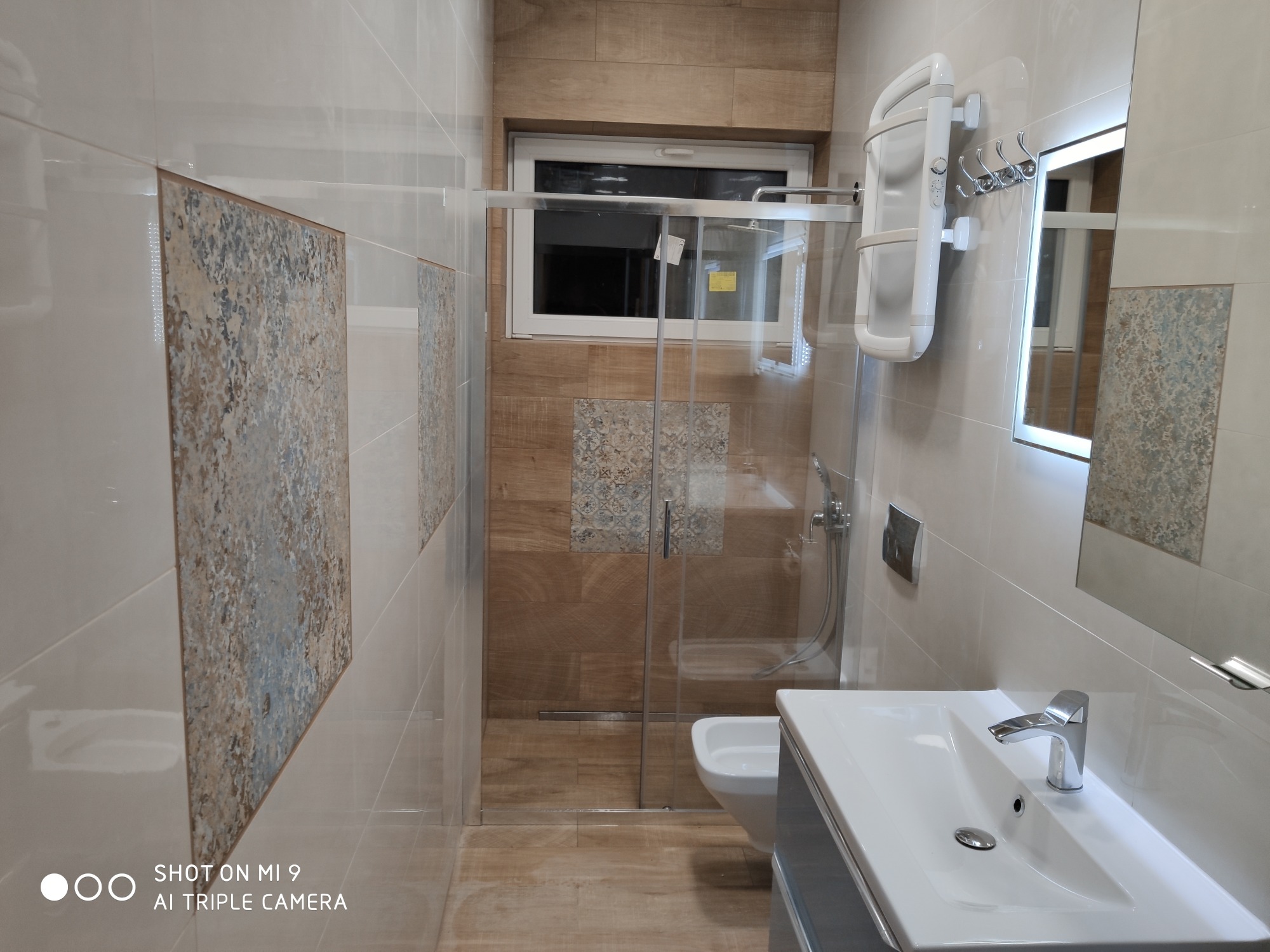 Nowoczesna łazienka po remoncie z kabiną prysznicową ze szklanymi drzwiami, umywalką z szafką w kolorze szarym, grzejnikiem drabinkowym i lustrem z podświetleniem LED.