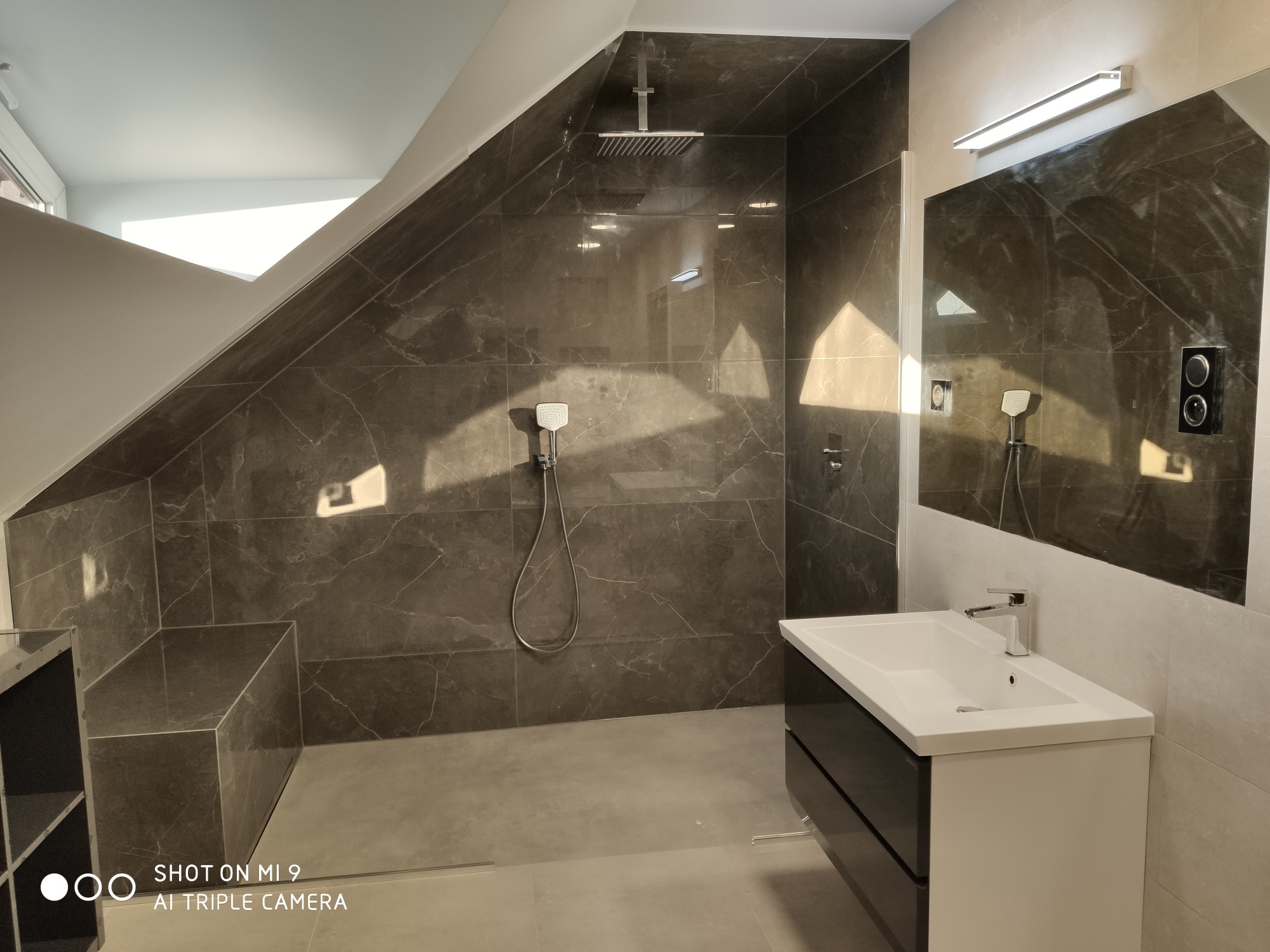 Wykończona łazienka na poddaszu z kabiną prysznicową typu walk-in wyłożoną ciemnymi płytkami imitującymi marmur, biała umywalka z czarnymi szufladami, oświetlenie LED nad lustrem.
