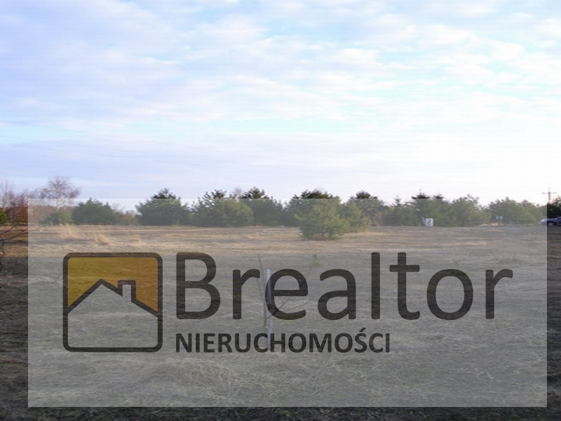 Działka budowlana z widocznymi drzewami w tle i logotypem agencji nieruchomości BRealtor na pierwszym planie.