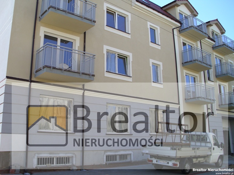 Nowoczesny budynek mieszkalny z balkonami i jasną elewacją, częściowo zasłonięty znakiem wodnym firmy 'BrealtoR Nieruchomości'.