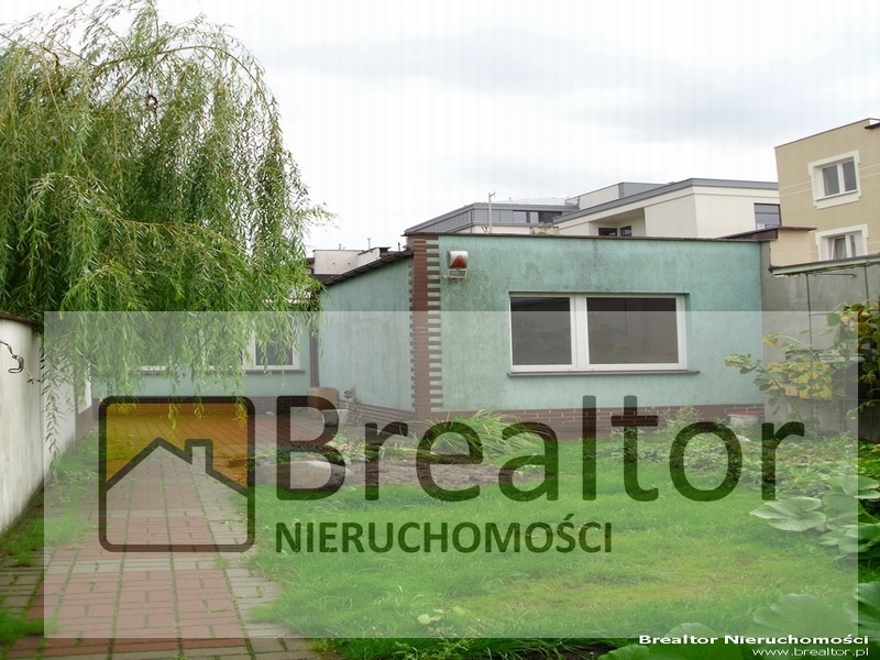 Niskie, turkusowe biuro z ceglanym kominem, widoczne zza drzewa i ścieżki z czerwonej kostki, z logo agencji nieruchomości na pierwszym planie.