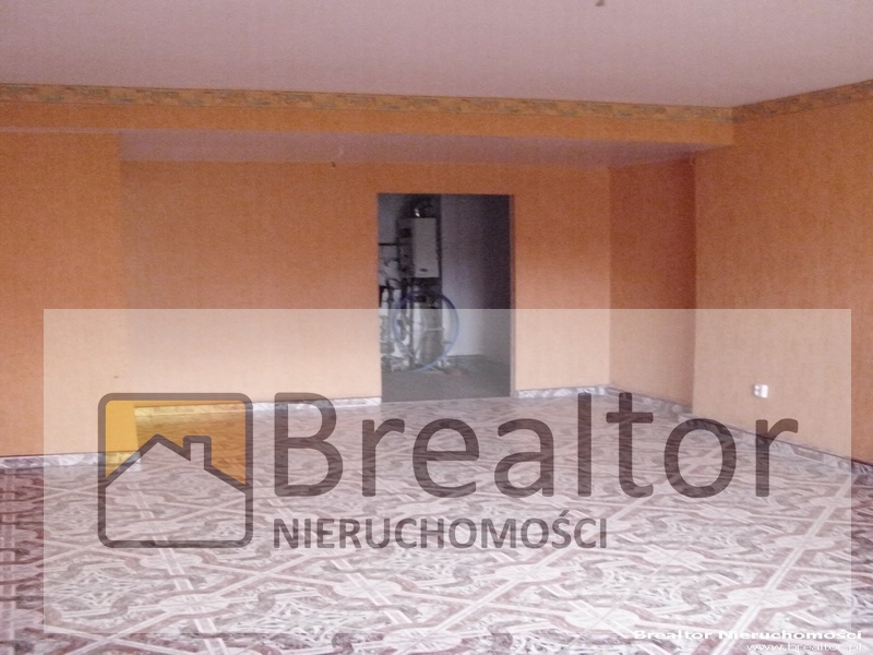 Puste pomieszczenie z pomarańczowymi ścianami, wzorzystą podłogą i otwartym przejściem do drugiego pokoju, gdzie widoczny jest rower. Na pierwszym planie logo agencji nieruchomości 'Brealtor'.