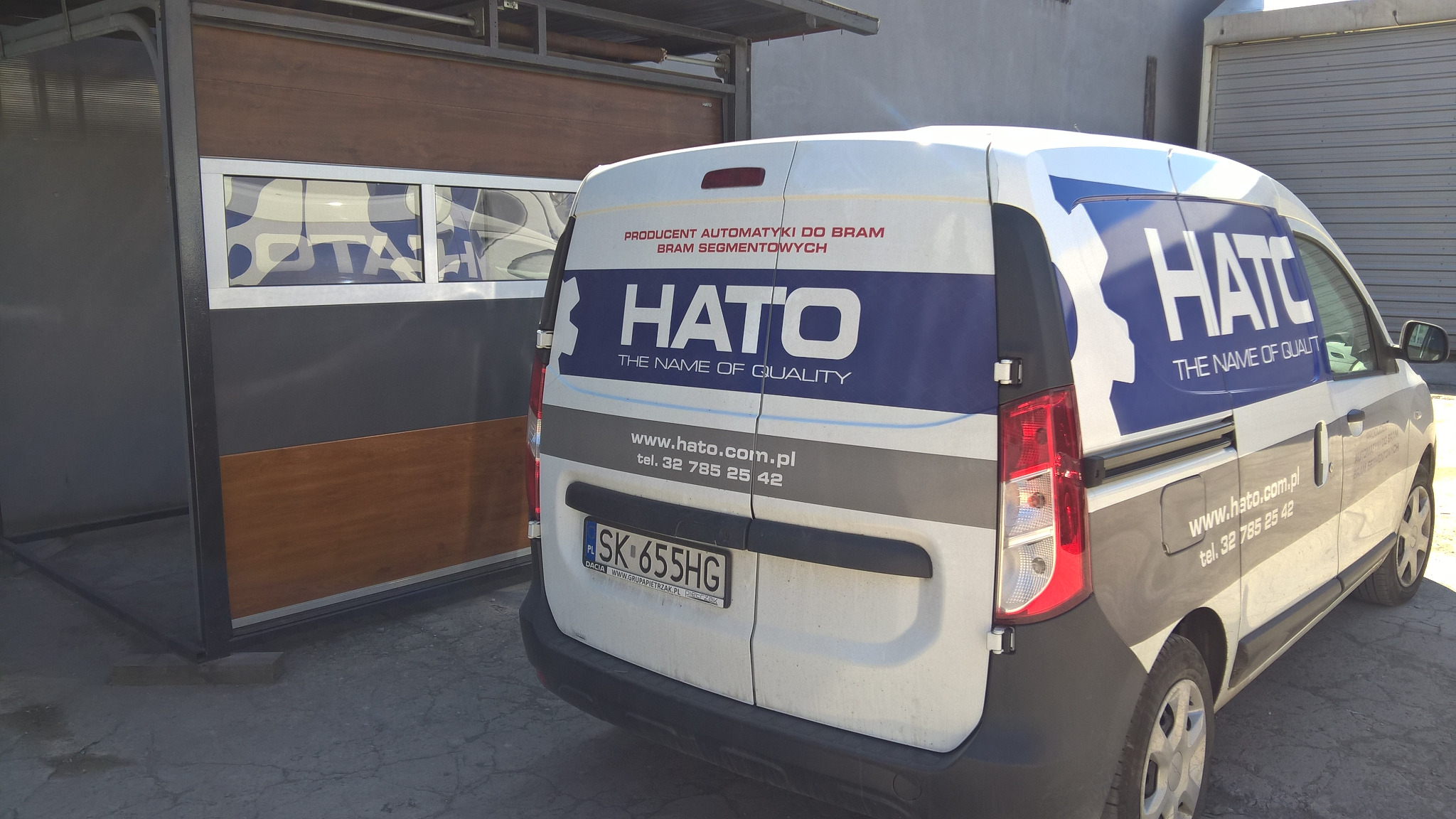 Samochód dostawczy z logo firmy HATO, producenta automatyki do bram segmentowych, zaparkowany obok ekspozycji bramy garażowej z oknami.