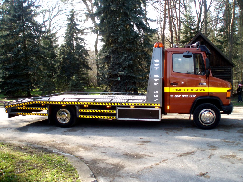 Pomarańczowa laweta Mercedes 811D z numerem telefonu '697 972 397' na tle drzew i drewnianej chaty.