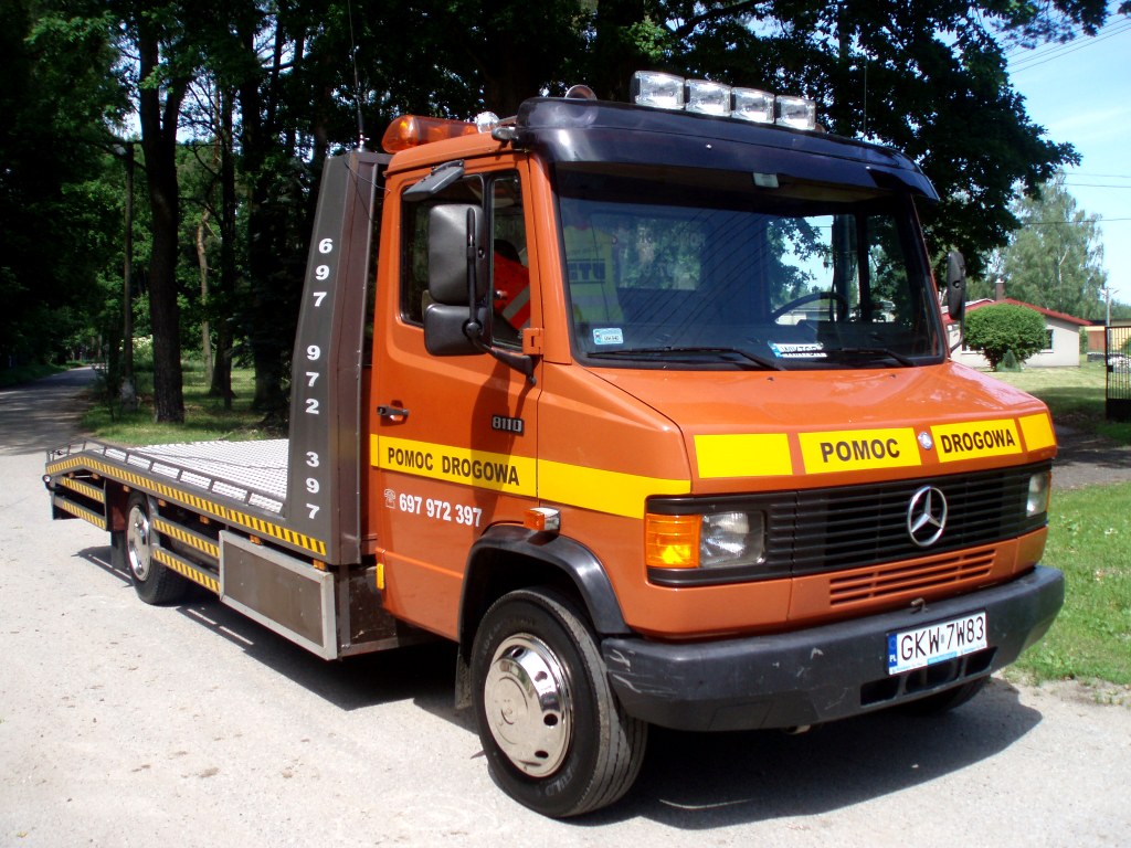 Pomarańczowa laweta Mercedes 811D z napisem 'POMOC DROGOWA' i numerem telefonu 697 972 397, zaparkowana na poboczu drogi z widocznymi drzewami w tle.