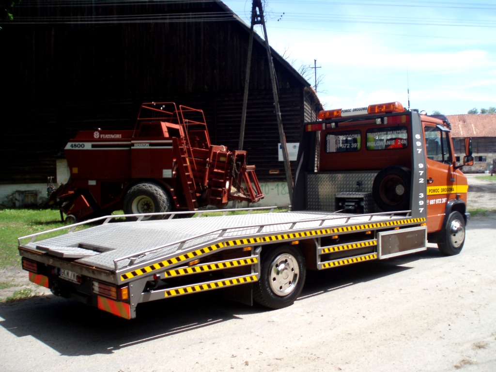 Pomarańczowa laweta z platformą załadunkową i żółto-czarnymi pasami, obok czerwona prasa rolnicza Fiatagri 4800, na tle drewnianego budynku gospodarczego.
