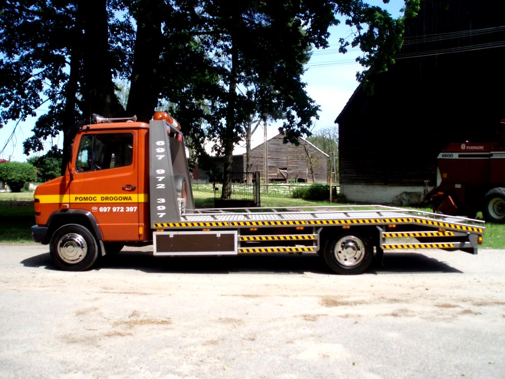 Pomarańczowy pojazd pomocy drogowej z napisem 'POMOC DROGOWA' i numerem telefonu, zaparkowany na asfaltowej drodze z wiejskimi budynkami w tle.