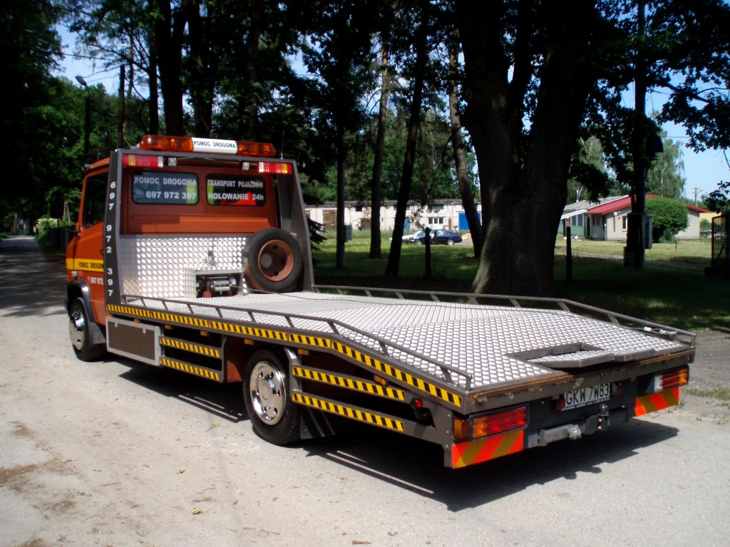 Pomarańczowa laweta z tabliczkami informacyjnymi i numerem telefonu, stojąca w Iławie na tle drzew i budynków, widok z tyłu.