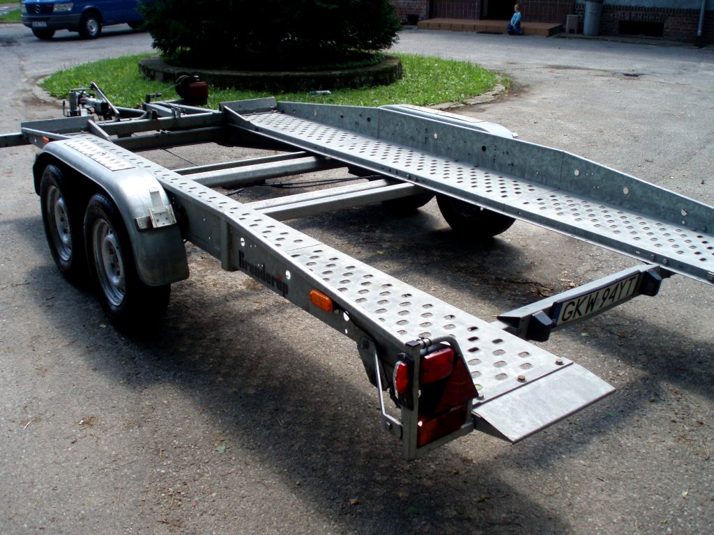 Pusta, dwuosiowa przyczepa samochodowa typu laweta z najazdami, zaparkowana na betonowym placu, widoczna tablica rejestracyjna.