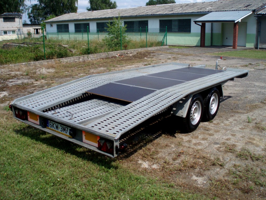 Pusta, dwuosiowa platforma lawety samochodowej z czarnymi panelami na środku, zaparkowana na trawie przed budynkiem gospodarczym.