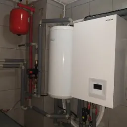 Biała jednostka Hitachi zamontowana na ścianie z szarymi płytkami, z widocznymi rurami, zaworami i czerwonym zbiornikiem wyrównawczym. Rury są zaizolowane szarą pianką.