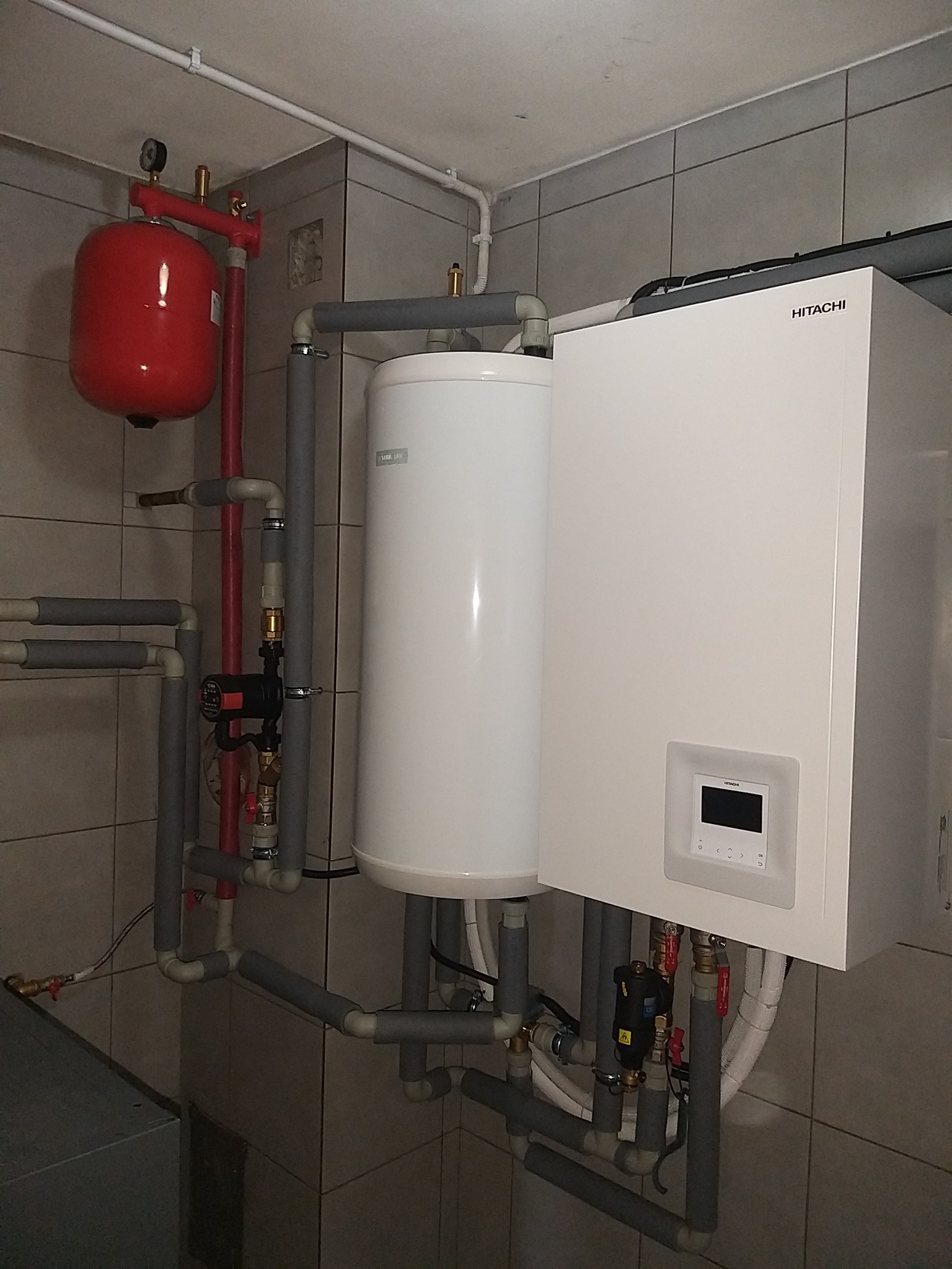 Biała jednostka Hitachi zamontowana na ścianie z szarymi płytkami, z widocznymi rurami, zaworami i czerwonym zbiornikiem wyrównawczym. Rury są zaizolowane szarą pianką.