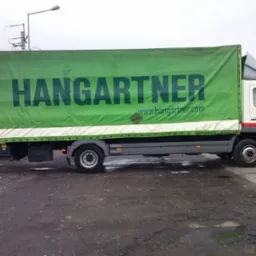 Ciężarówka z zieloną plandeką firmy Hangartner zaparkowana na mokrym placu.
