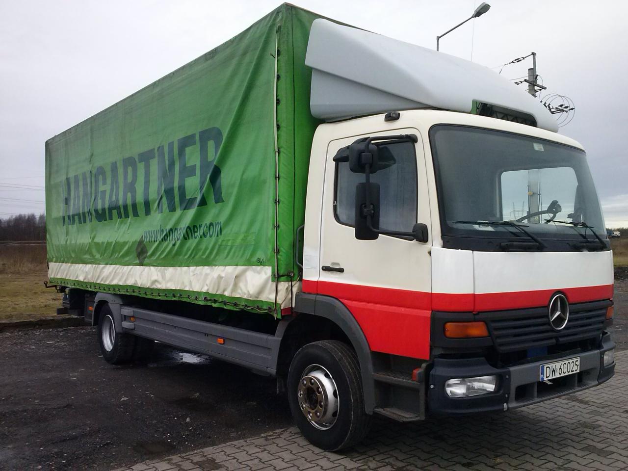 Ciężarówka Mercedes z zieloną plandeką z napisem 'HANGARTNER' na parkingu z kostki brukowej, widok z boku.
