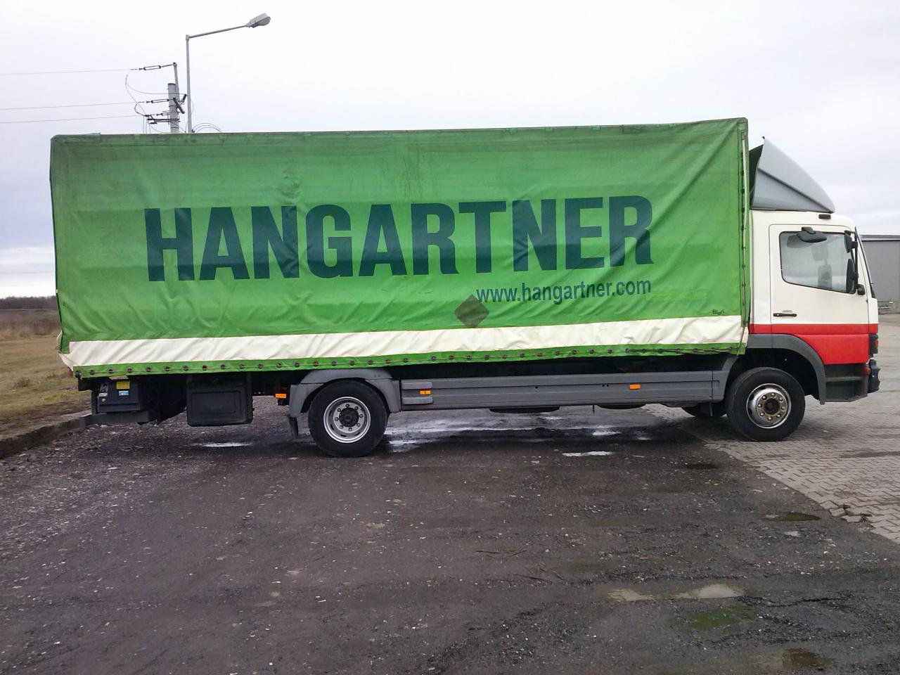 Ciężarówka z zieloną plandeką firmy Hangartner zaparkowana na mokrym placu.