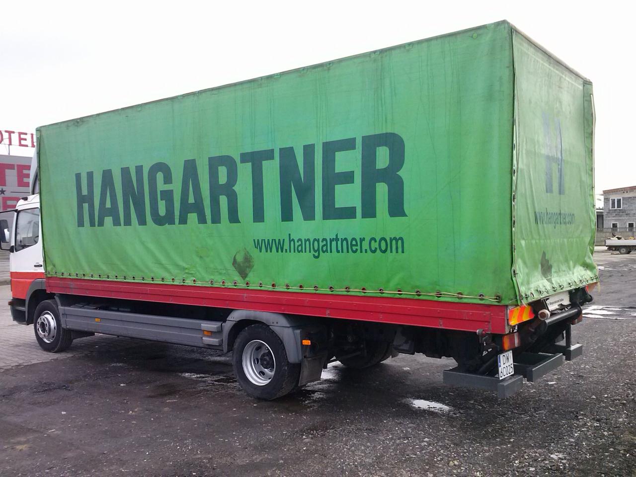 Ciężarówka z zieloną plandeką i napisem HANGARTNER oraz adresem strony internetowej, zaparkowana na placu z widocznym hotelem w tle.