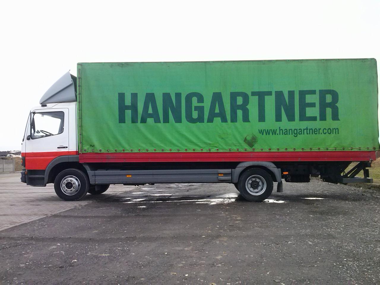 Ciężarówka z zieloną plandeką z napisem HANGARTNER i adresem strony internetowej, stojąca na mokrym placu.