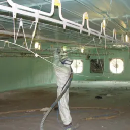 Pracownik w kombinezonie ochronnym natryskuje pianę poliuretanową na ściany i sufit hali przemysłowej, widoczne wentylatory i oświetlenie pod sufitem.