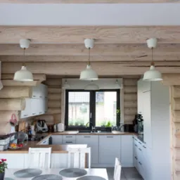 Jasna kuchnia w domu z bali, widoczne drewniane ściany, białe szafki kuchenne, drewniany blat, czarny zlew i czarne okno, trzy białe lampy wiszące nad stołem.
