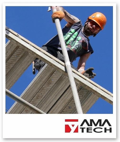 Pracownik w pomarańczowym kasku i okularach ochronnych montuje metalowe rusztowanie na tle błękitnego nieba, widok z dołu.