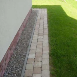 Chodnik z kostki brukowej przylegający do budynku, oddzielony od trawnika, z wąskim pasem żwiru wzdłuż ściany.