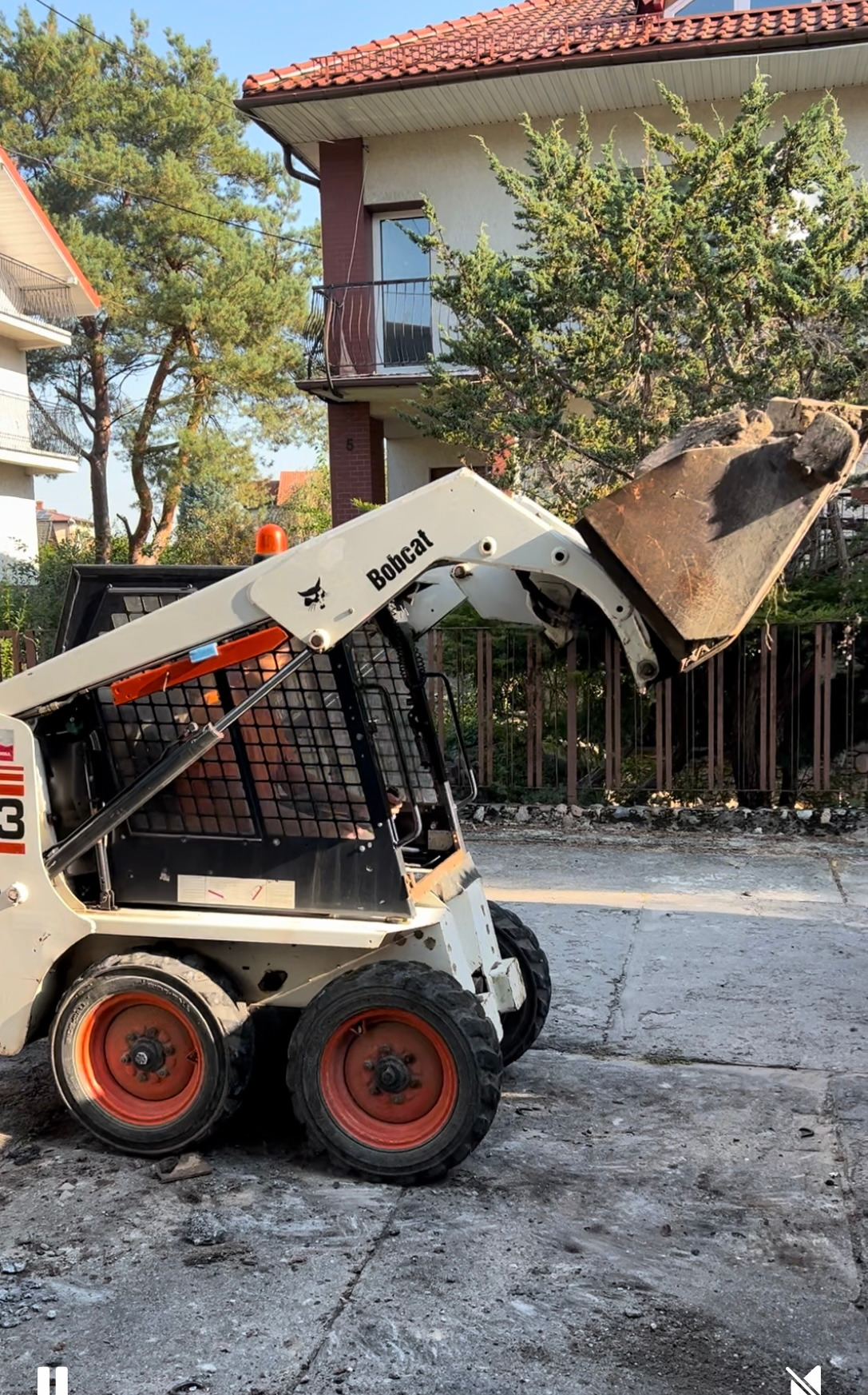 Bobcat z pełną łyżką gruzu podczas rozbiórki w Olsztynie; widoczne fragmenty zdemontowanego ogrodzenia.