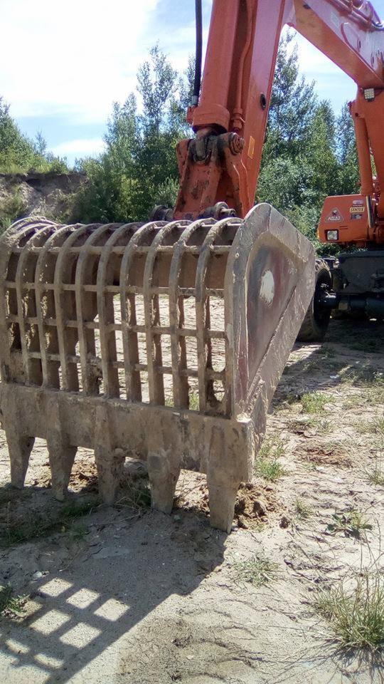 Pomarańczowa koparka z kratownicową łyżką, stojąca na piaszczystym terenie z zielenią w tle.
