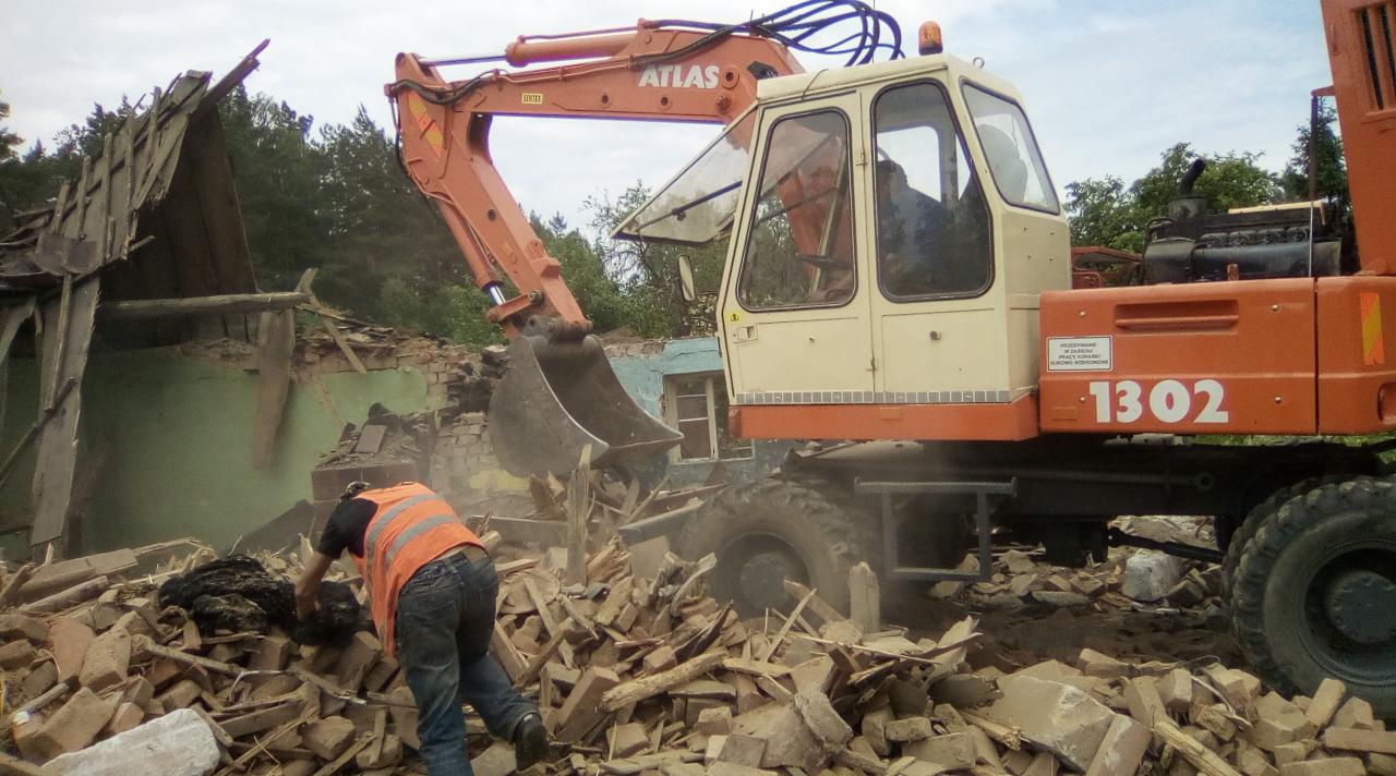 Pomarańczowo-beżowa koparka Atlas w trakcie rozbiórki budynku. Operator w kabinie i pracownik w kamizelce odblaskowej wśród gruzu. Częściowo zburzona, zielona ściana w tle.
