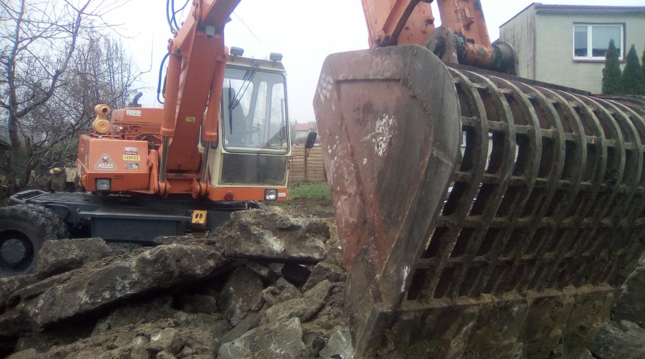 Pomarańczowa koparka kołowa Atlas Sentex z ażurową łyżką przesiewającą, pracująca na gruzowisku z fragmentami betonu i ziemi, w tle budynek mieszkalny i drzewa bez liści.