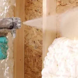 Aplikacja pianki poliuretanowej natryskowej na drewnianą konstrukcję szkieletową ściany, widoczne narzędzie aplikacyjne z niebieskimi przewodami i dłoń w zielonej rękawicy ochronnej.