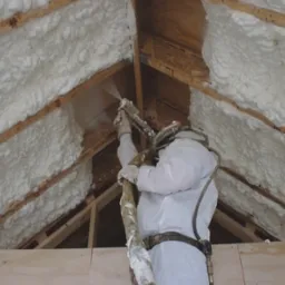 Pracownik w kombinezonie ochronnym natryskuje piankę izolacyjną na drewnianą konstrukcję dachu poddasza, widoczne świeżo nałożone warstwy pianki.