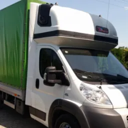 Biały bus z zieloną plandeką na tle pola i drzew, zaparkowany na brukowanej nawierzchni.