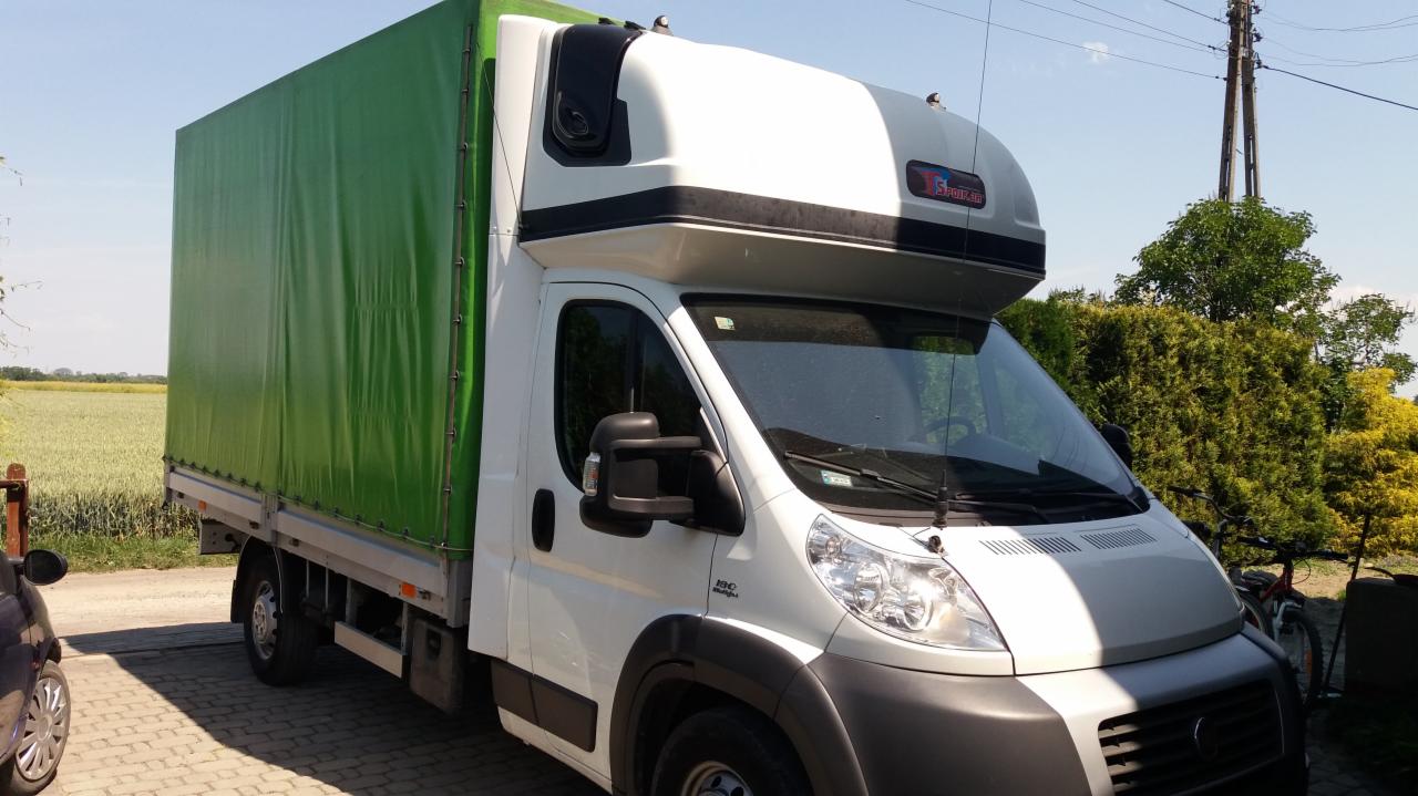 Biały bus z zieloną plandeką na tle pola i drzew, zaparkowany na brukowanej nawierzchni.