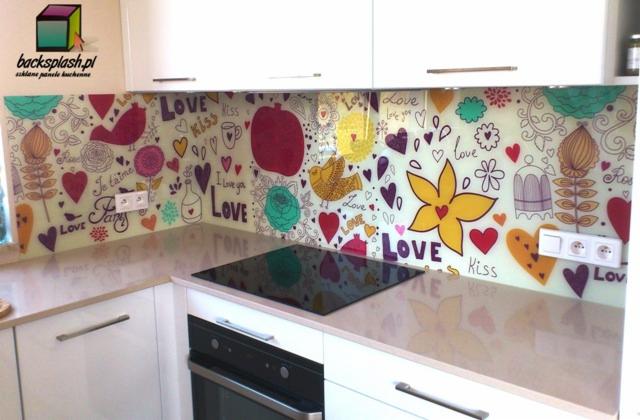 Szklany panel kuchenny z kolorowym, walentynkowym wzorem, zamontowany nad płytą grzewczą. Białe szafki kuchenne w tle, widoczne gniazdka elektryczne.