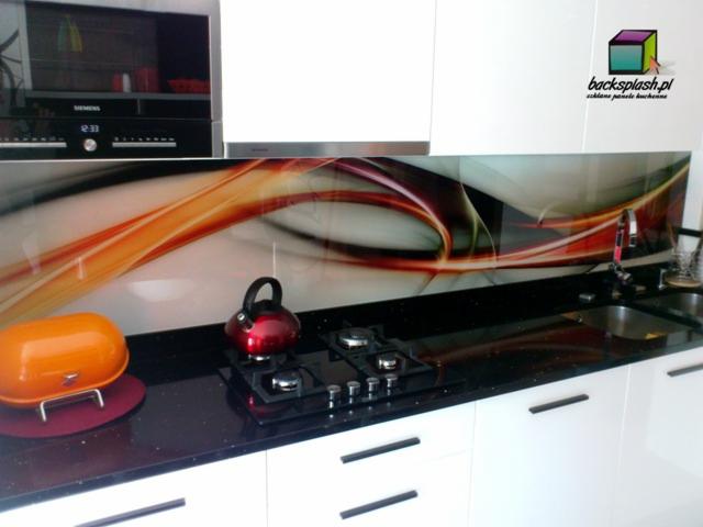 Biała kuchnia z czarnym blatem i panelem szklanym z abstrakcyjnym wzorem w odcieniach czerwieni i pomarańczy. Na blacie czajnik i chlebak.