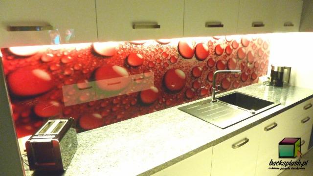 Kuchnia z czerwonym panelem szklanym z motywem kropli, podświetlonym od góry. Widoczny zlew, bateria i toster na blacie. Nowoczesny design.