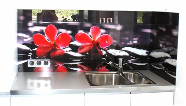 Szklana ściana nad blatem kuchennym z motywem kwiatów i kamieni, odbijająca się w blacie. Nowoczesny design, srebrny zlew. Widoczne białe szafki.