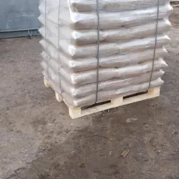 Paleta z workami pelletu owinięta folią i zabezpieczona taśmami, na ziemi przed budynkiem.