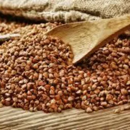 Kupa kaszy gryczanej na drewnianym stole, częściowo przykryta jutowym workiem, z drewnianą łyżką zanurzoną w kaszy.