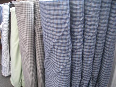 Rzędy pionowo ustawionych rolek tkanin o różnych wzorach: paski, kratka, gładka z widocznymi krawędziami i fakturą materiałów.