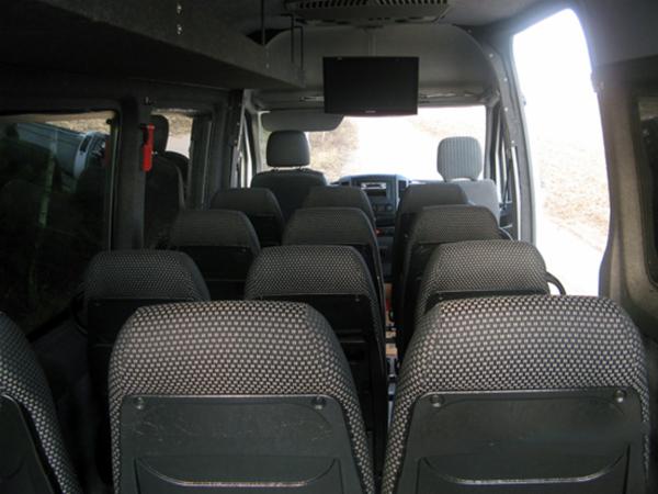 Wnętrze busa pasażerskiego z rzędami foteli z czarną tapicerką w kratkę i zamontowanym ekranem pod sufitem, widok z tyłu pojazdu.