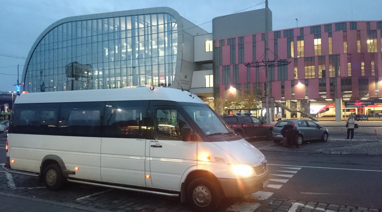 Biały bus przed budynkiem dworca Poznań Główny. W tle widoczny pociąg i osoby czekające na peronie. Oświetlenie uliczne włączone, zmierzch.