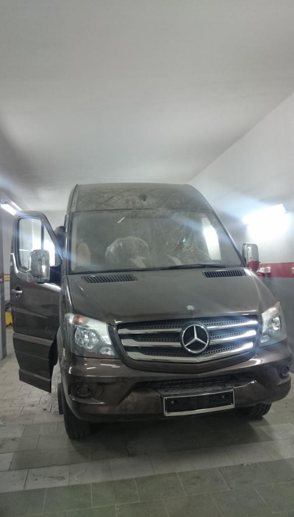 Brązowy Mercedes Sprinter z otwartymi drzwiami w garażu. Widoczne detale: chromowany grill, reflektory i logo marki. Auto sprawia wrażenie używanego.