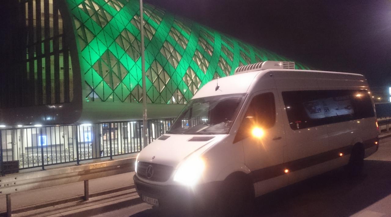 Biały bus Mercedes-Benz Sprinter z klimatyzacją na dachu, zaparkowany nocą przed nowoczesnym budynkiem z zielonym, geometrycznym oświetleniem.