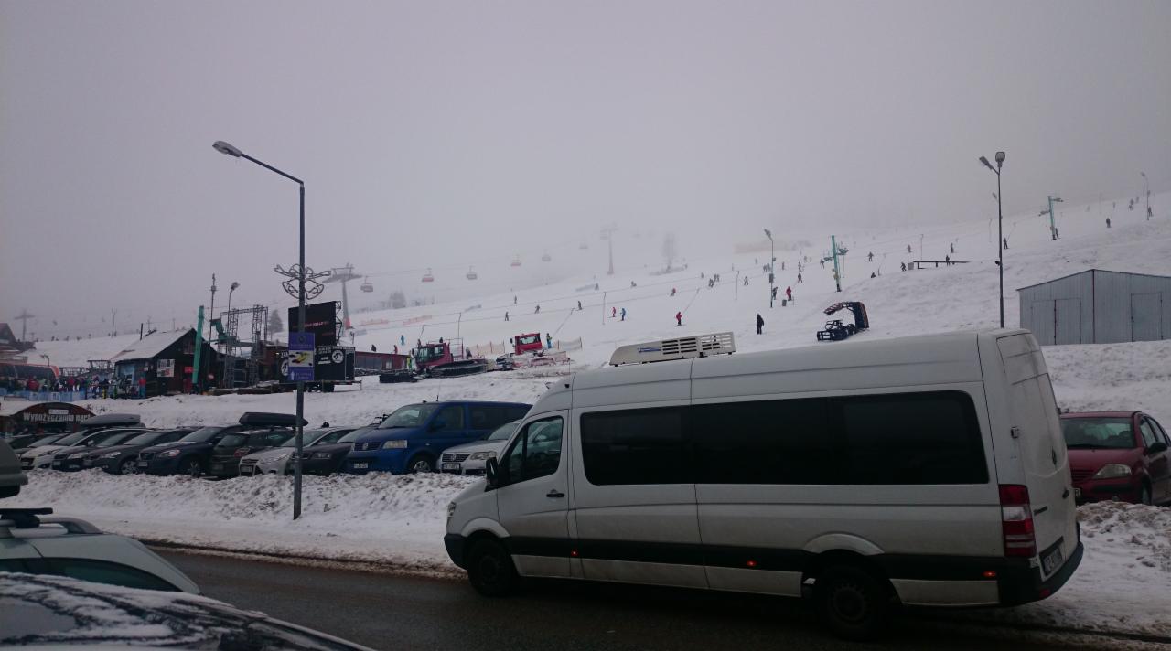 Biały bus na parkingu przy stoku narciarskim w zimowej scenerii. W tle widać narciarzy i wyciągi. Pochmurny dzień.
