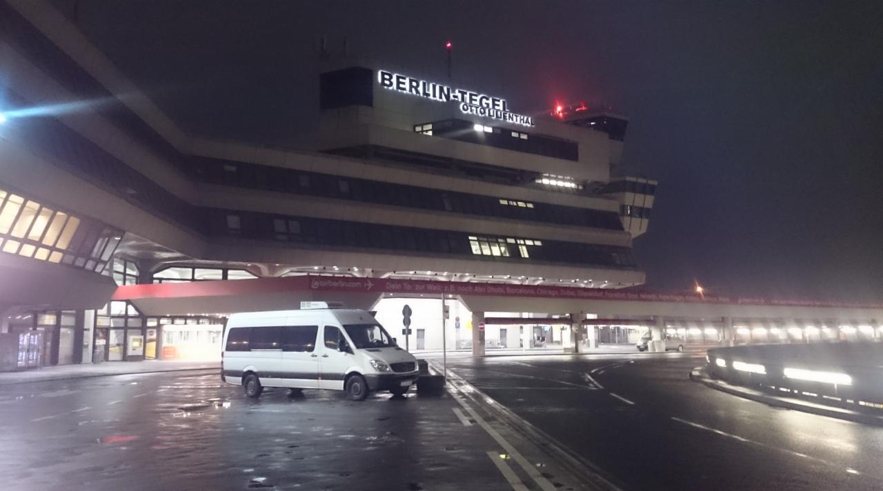 Biały bus przed lotniskiem Berlin-Tegel nocą, mokra nawierzchnia, światła odbijają się w kałużach, widoczny budynek lotniska z napisem.
